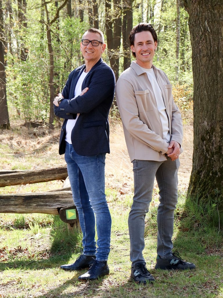 Two men smiling at camera standing in a forest with tall trees and fallen logs behind them
